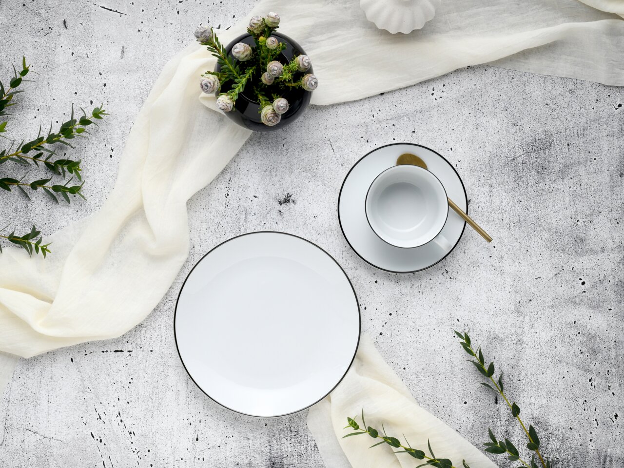 Kaffeeservice Luna Black L, bestehend aus einem Teller und einer Tasse mit Untertasse, von oben fotografiert. Elegantes Design mit schwarzem Rand auf weißem Porzellan, dekoriert mit einem goldenen Löffel und umgeben von grünen Zweigen und einem weißen Tuc