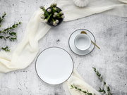 Kaffeeservice Luna Black L, bestehend aus einem Teller und einer Tasse mit Untertasse, von oben fotografiert. Elegantes Design mit schwarzem Rand auf weißem Porzellan, dekoriert mit einem goldenen Löffel und umgeben von grünen Zweigen und einem weißen Tuc