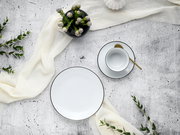 Kaffeeservice Luna Black L, bestehend aus einem Teller und einer Tasse mit Untertasse, von oben fotografiert. Elegantes Design mit schwarzem Rand auf weißem Porzellan, dekoriert mit einem goldenen Löffel und umgeben von grünen Zweigen und einem weißen Tuc