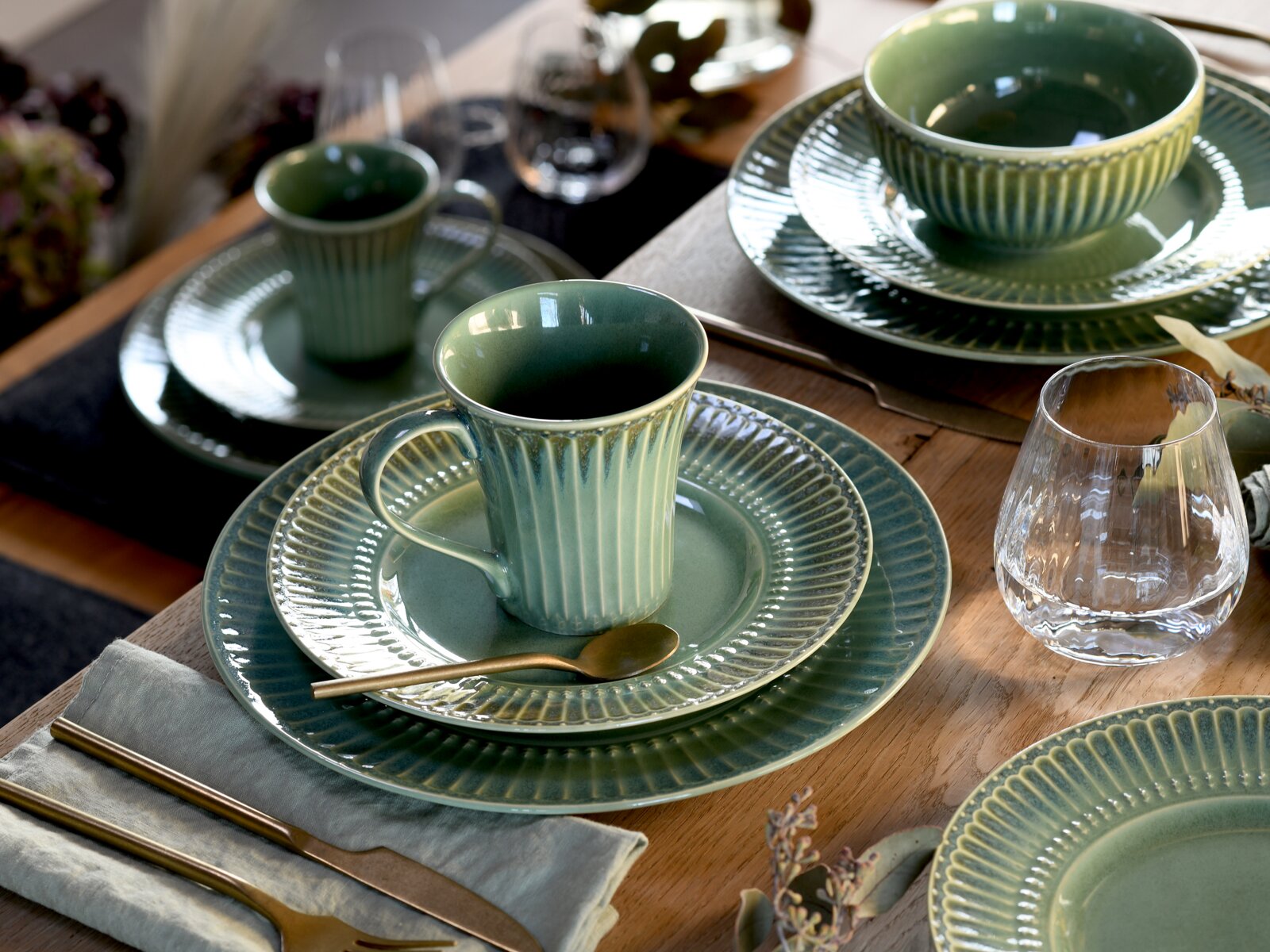 Schräg von oben fotografiertes Tischgedeck mit grün glasiertem, geripptem Keramikgeschirr: Tasse auf Untertasse und Speiseteller sowie passende Schalen, dazu Glas und goldfarbenes Besteck auf Holztisch.