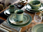 Schräg von oben fotografiertes Tischgedeck mit grün glasiertem, geripptem Keramikgeschirr: Tasse auf Untertasse und Speiseteller sowie passende Schalen, dazu Glas und goldfarbenes Besteck auf Holztisch.