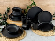 Schwarzes Kombiservice 16-tlg aus Keramik, bestehend aus Tellern, Schalen und Bechern, auf einem Tisch mit Holz- und Pflanzenakzenten, aus frontaler Perspektive fotografiert.