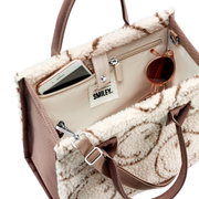 Schräg von oben in die offene, cremefarbene Teddy-Handtasche mit braunen Tragegriffen und Smiley-Label, innen mit Reißverschlussfach; Smartphone und Sonnenbrille sind sichtbar.