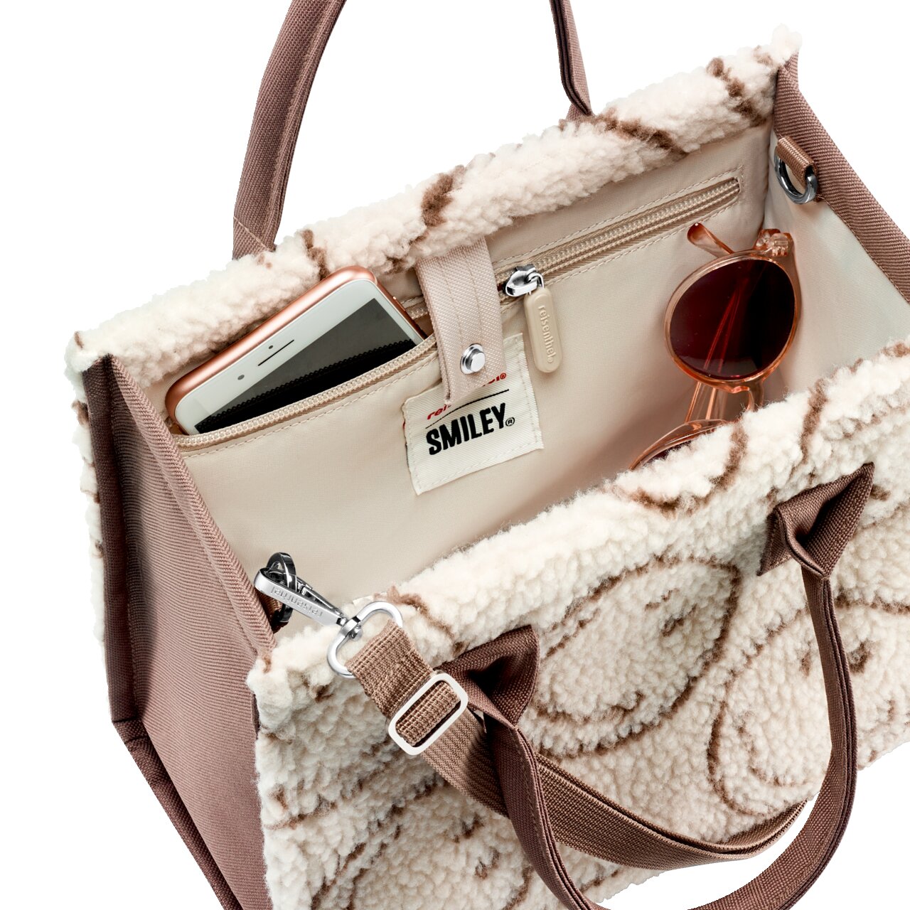 Schräg von oben in die offene, cremefarbene Teddy-Handtasche mit braunen Tragegriffen und Smiley-Label, innen mit Reißverschlussfach; Smartphone und Sonnenbrille sind sichtbar.