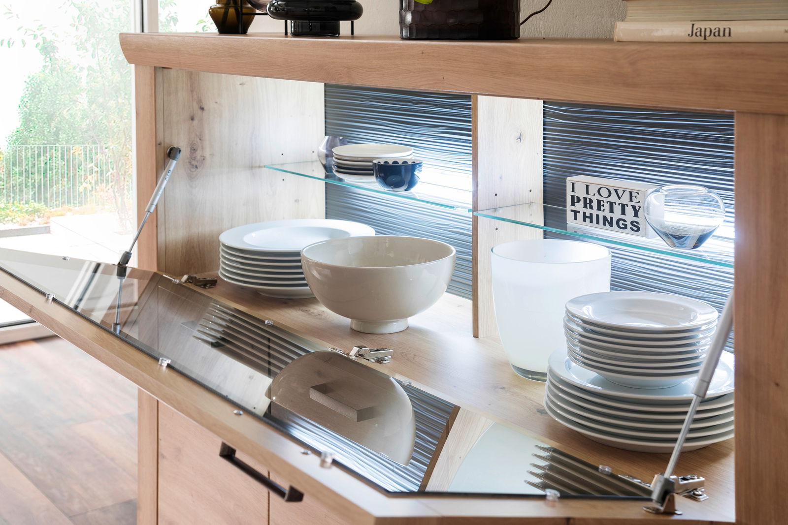 Highboard mit geöffneter Glastür, gefüllt mit Geschirr und Dekoration, aus der Perspektive von vorne.