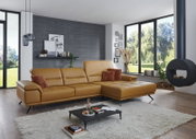 Modernes Wohnzimmersetting mit cognacfarbenem Leder-Ecksofa mit rechter Ottomane/Longchair, gesteppten Sitzflächen und schwarzen Metallfüßen; Perspektive: frontale, leicht seitliche Ansicht von links auf das Sofa.