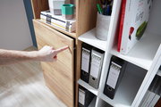 Nahaufnahme eines Aktenschranks mit Holz- und weißen Elementen, in dem Ordner und Bücher aufbewahrt werden. Eine Hand zeigt auf die Schranktür. Perspektive von der Seite.