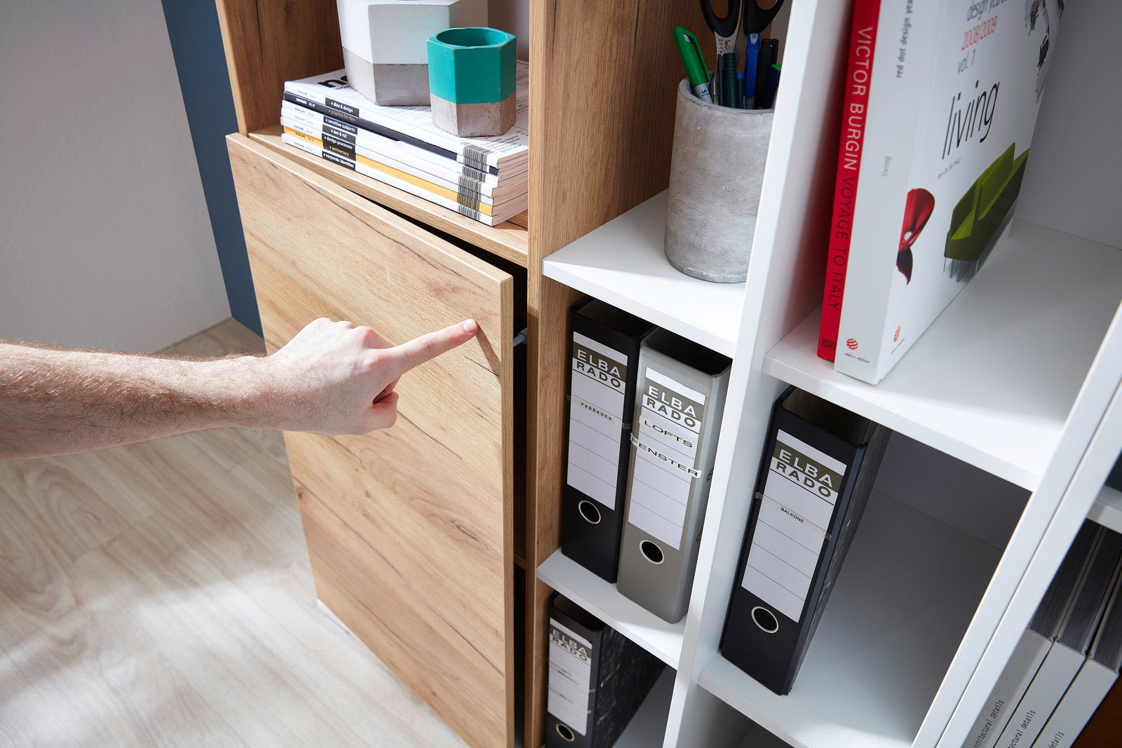 Nahaufnahme eines Aktenschranks mit Holz- und weißen Elementen, in dem Ordner und Bücher aufbewahrt werden. Eine Hand zeigt auf die Schranktür. Perspektive von der Seite.