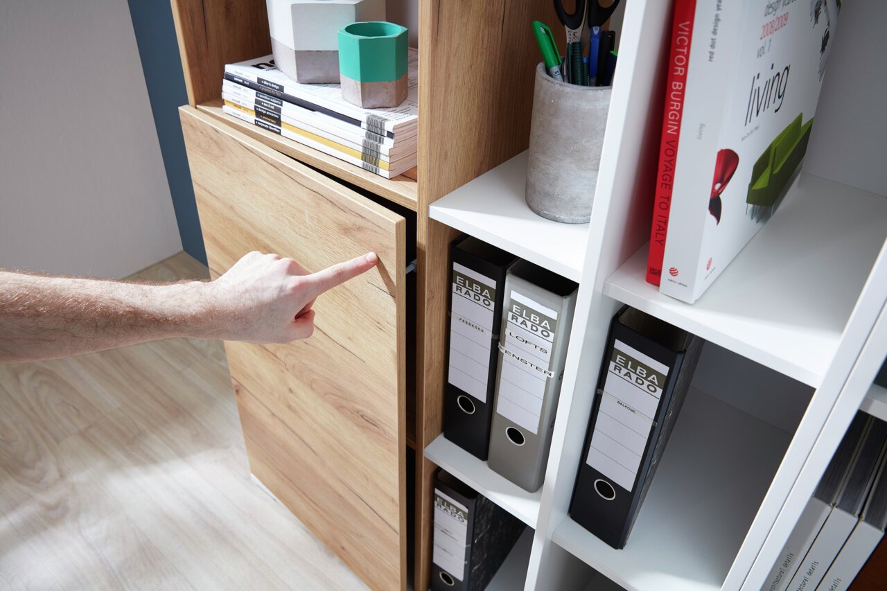 Nahaufnahme eines Aktenschranks mit Holz- und weißen Elementen, in dem Ordner und Bücher aufbewahrt werden. Eine Hand zeigt auf die Schranktür. Perspektive von der Seite.