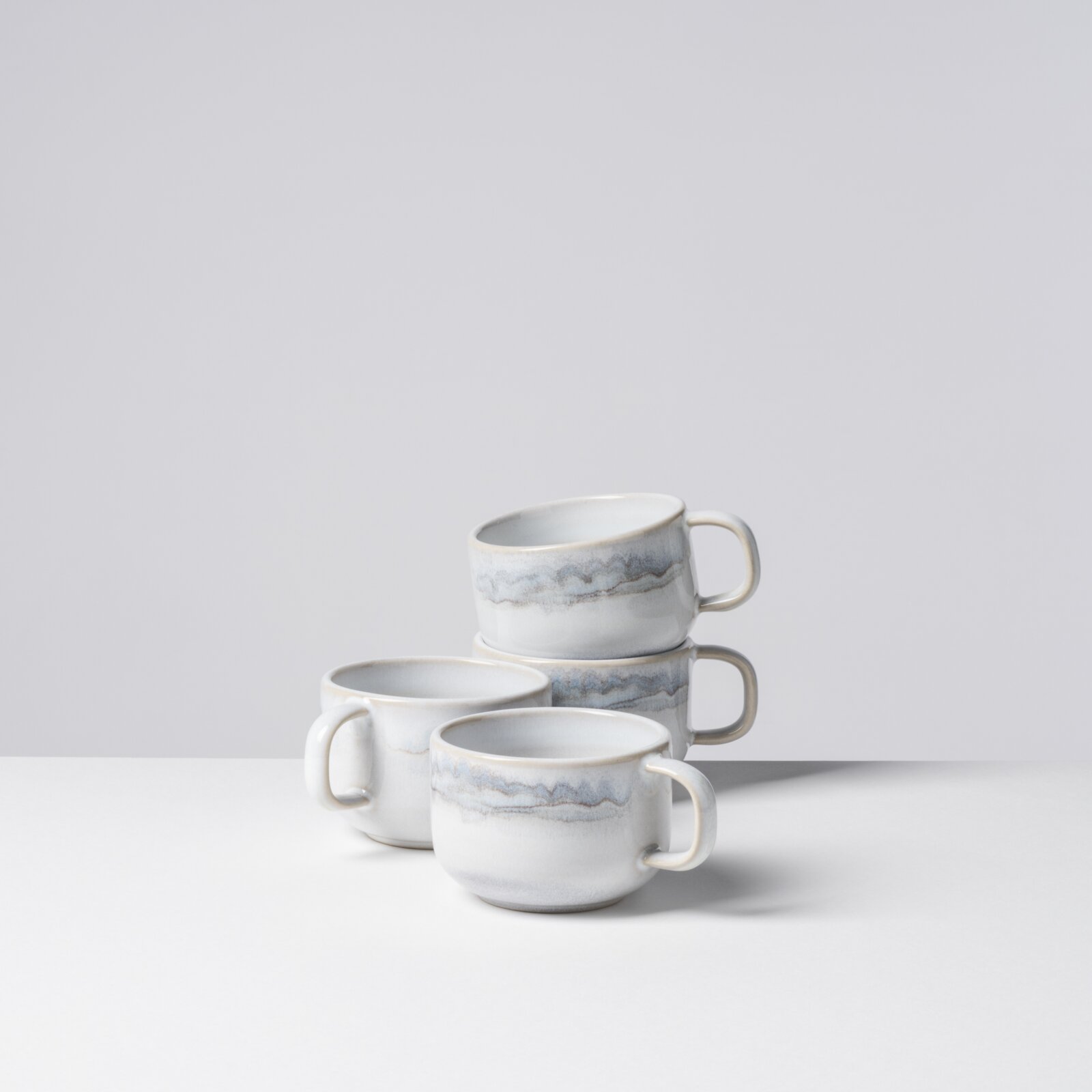 Drei stapelbare Keramik-Kaffeetassen in Weiß mit graublauer Marmorierung und Henkel, auf weißem Untergrund vor hellem Hintergrund, fotografiert in leichter Seitenansicht von schräg oben.