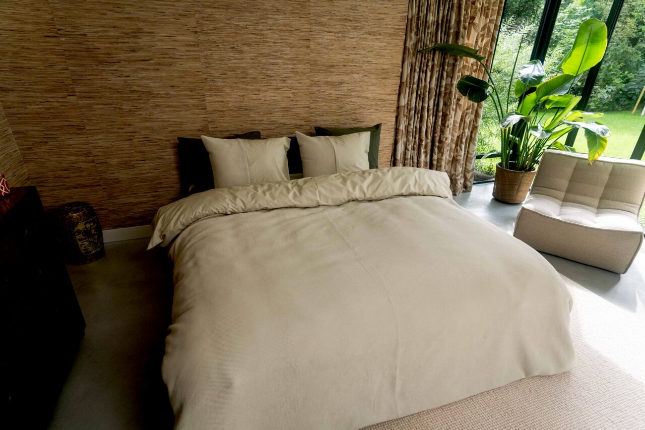 Kayori Bettwäsche ERIMO Gemütliches Schlafzimmer mit beiger Bettwäsche auf einem Doppelbett, umgeben von natürlichen Elementen und einem großen Fenster mit Blick ins Grüne. Perspektive von vorne.
