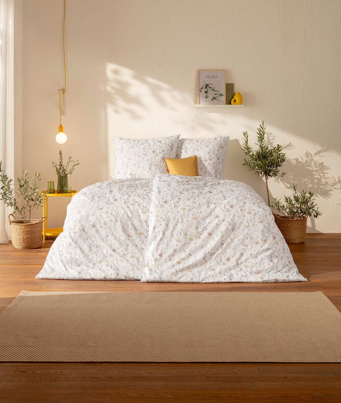 Estella Mako-Satin Bettwäsche 135/200cm CELESTE Frontalansicht: Weißer Bettbezug mit zartem Blumenmuster und passenden Kissen in hellem, sonnigem Schlafzimmer mit gelbem Akzentkissen, Hängeleuchte und Pflanzen.