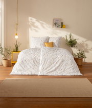Frontalansicht: Weißer Bettbezug mit zartem Blumenmuster und passenden Kissen in hellem, sonnigem Schlafzimmer mit gelbem Akzentkissen, Hängeleuchte und Pflanzen.