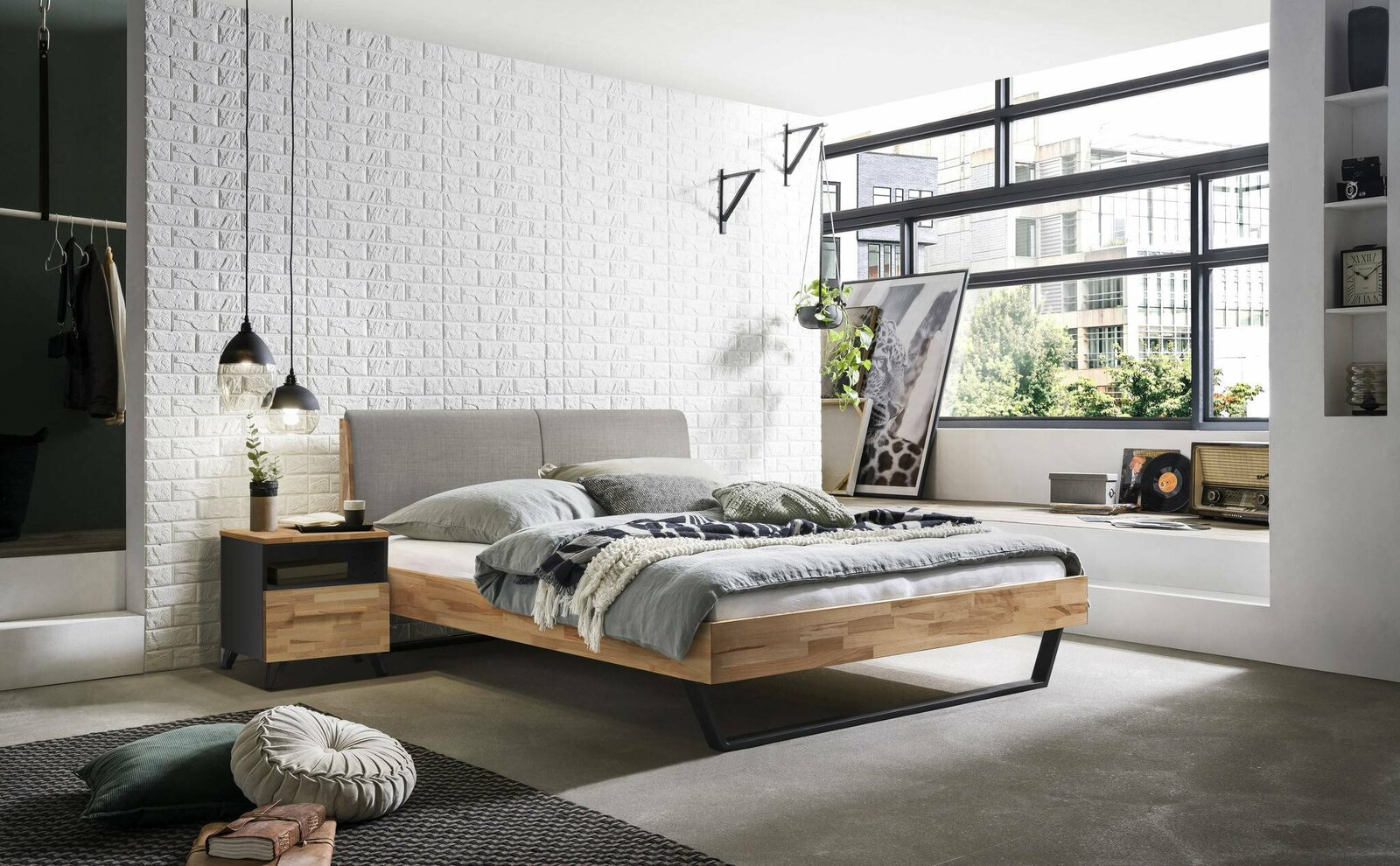 Modernes Bettgestell aus Holz mit grauem Kopfteil, seitliche Perspektive, in einem stilvoll eingerichteten Schlafzimmer mit großen Fenstern und dekorativen Elementen.
