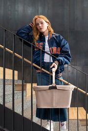 Eine Frau in einer blauen Jacke steht auf einer Treppe und hält einen beigen Einkaufskorb mit Griff. Der Korb wird von vorne gezeigt.