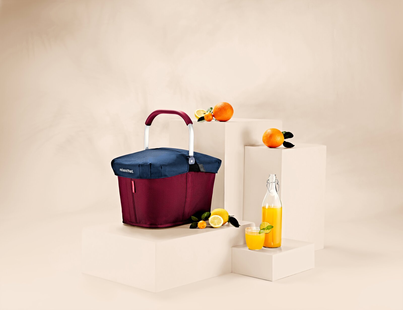 Reisenthel Carrybag-hoes donkerblauw Draagtas met donkerblauwe hoes, omringd door sinaasappels en citroenen, op witte sokkels. Perspectief: zijaanzicht.