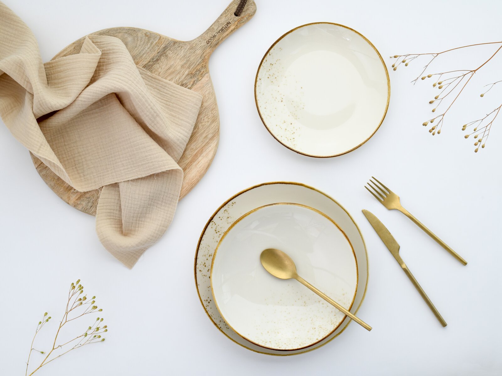 Draufsicht (Flatlay) auf zwei cremeweiße Keramikschalen mit goldfarbenem Rand und Sprenkeln, goldfarbenem Besteck, Holzbrett mit beigefarbenem Stoff und Trockenblumen auf weißem Hintergrund