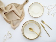 Draufsicht (Flatlay) auf zwei cremeweiße Keramikschalen mit goldfarbenem Rand und Sprenkeln, goldfarbenem Besteck, Holzbrett mit beigefarbenem Stoff und Trockenblumen auf weißem Hintergrund