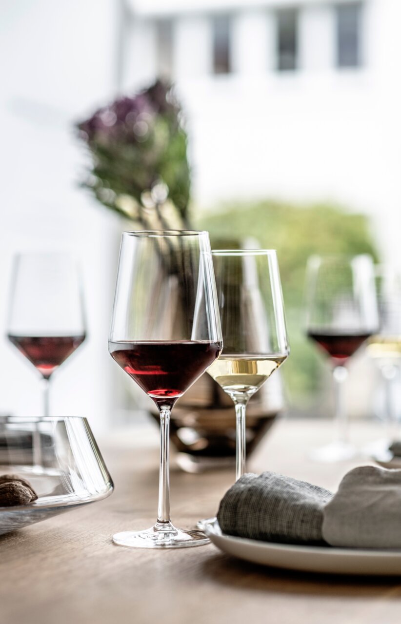 Een Cabernet-rodewijnglas uit de Pure-serie, gevuld met rode wijn, op een gedekte tafel in een moderne, lichte omgeving. Gezien vanuit een schuine hoek van voren.