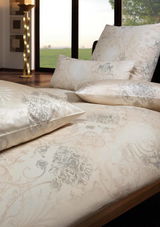Luxuriöse Bettwäsche in Creme- und Grautönen mit floralem Muster, auf einem Bett arrangiert, mit Blick auf ein Fenster mit Landschaft im Hintergrund.