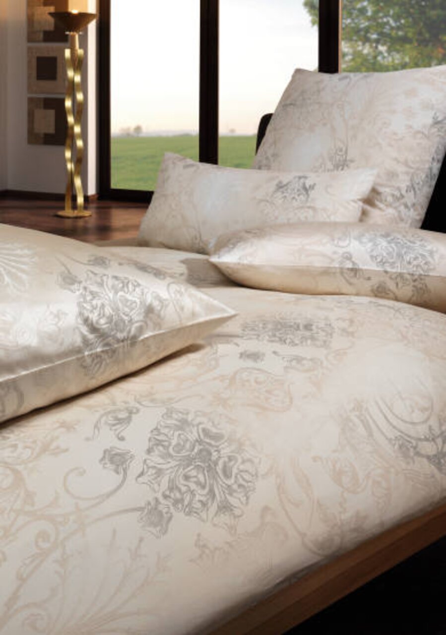 Luxuriöse Bettwäsche in Creme- und Grautönen mit floralem Muster, auf einem Bett arrangiert, mit Blick auf ein Fenster mit Landschaft im Hintergrund.
