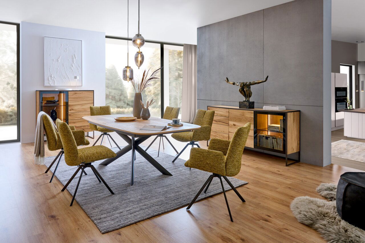 Modernes Esszimmer mit einem eleganten Holztisch und stilvollen Stühlen in einem hellen Raum. Im Hintergrund sind ein Highboard und ein Sideboard aus Holz mit Glasdetails zu sehen. Die Perspektive zeigt den Raum von der Seite, mit Blick auf den Tisch und