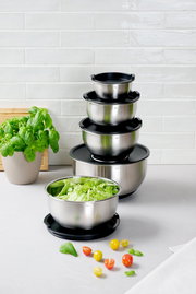 Ein Set aus fünf stapelbaren Rührschüsseln aus Edelstahl mit schwarzen Deckeln, präsentiert auf einem Tisch mit frischem Salat und Kirschtomaten. Perspektive von vorne.