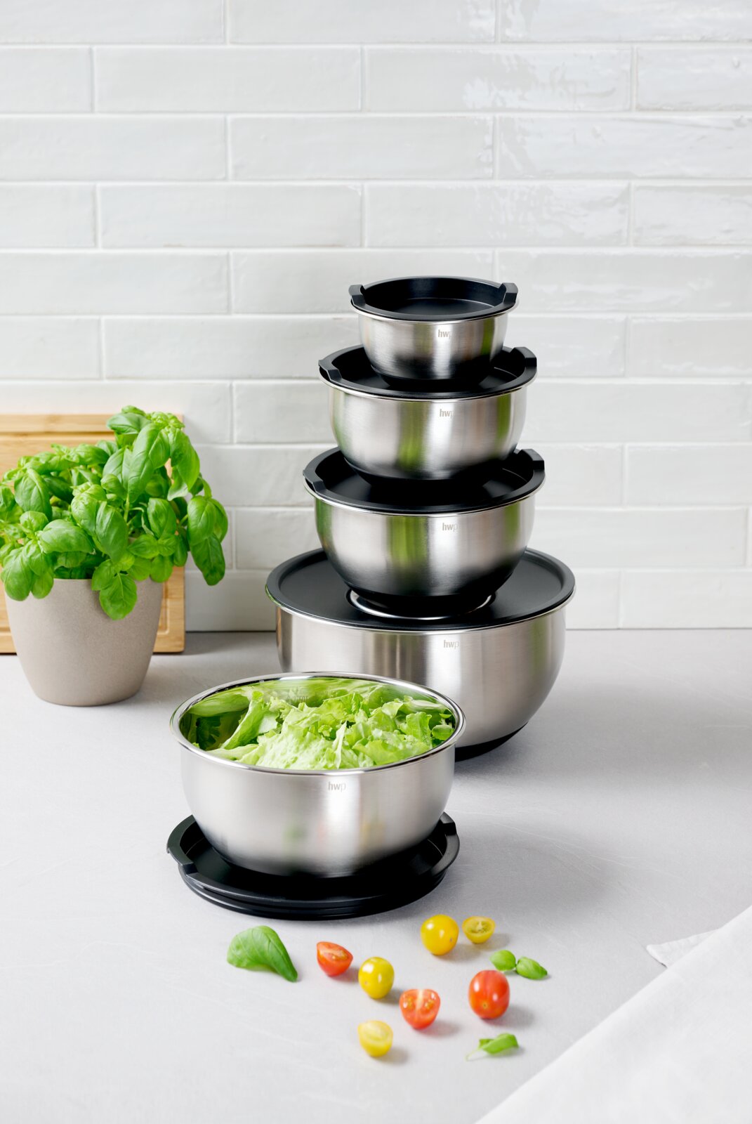 Homeware Rührschüssel-Set 8tlg. Ein Set aus fünf stapelbaren Rührschüsseln aus Edelstahl mit schwarzen Deckeln, präsentiert auf einem Tisch mit frischem Salat und Kirschtomaten. Perspektive von vorne.