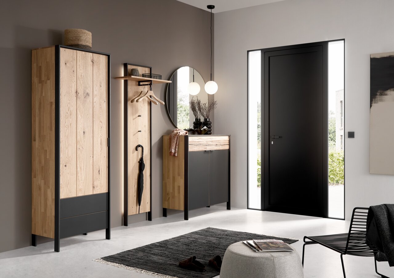 Moderne Garderobeneinrichtung mit Holz- und Metallelementen, bestehend aus einem hohen Schrank, einem Garderobenpaneel mit Haken und einem runden Spiegel, aus der Perspektive des Eingangsbereichs betrachtet.