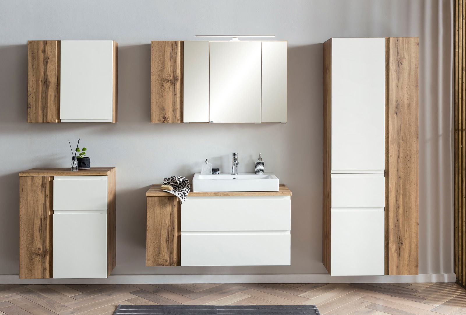 Moderne Badezimmermöbel in Holzoptik mit weißen Fronten, bestehend aus Hochschrank, Waschbeckenunterschrank und Hängeschränken, frontal betrachtet.