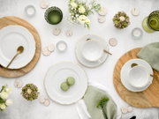 Draufsicht auf eine stilvolle Tischdekoration mit weißem Porzellan-Geschirr mit goldfarbenem Rand, Tassen und Tellern auf Holzplatten, grünen Gläsern, Teelichtern, Blumenarrangements und zwei grünen Macarons auf hellem Hintergrund.