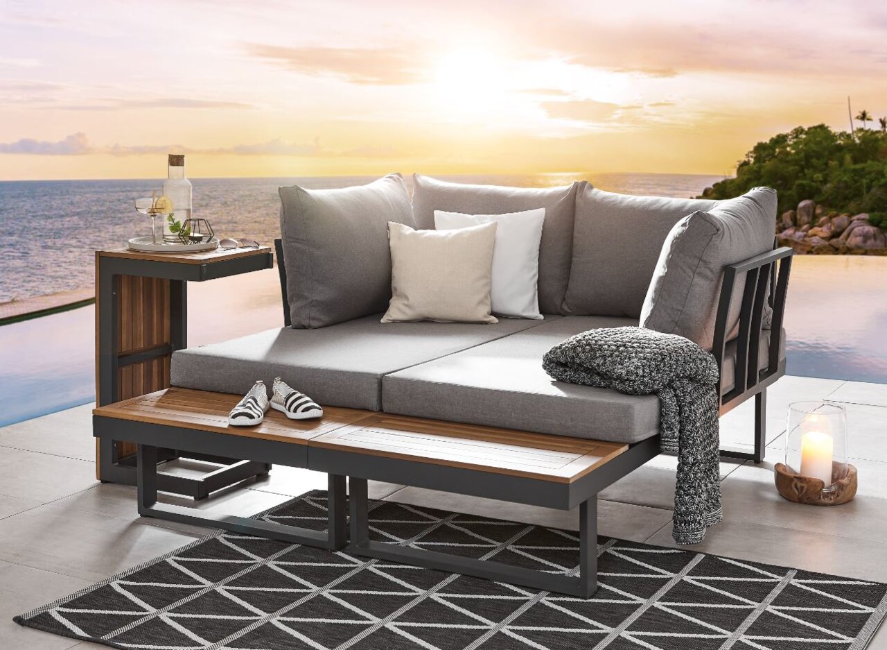 Gartenlounge mit grauen Kissen und Holzdetails, seitliche Perspektive, im Hintergrund Sonnenuntergang über dem Meer