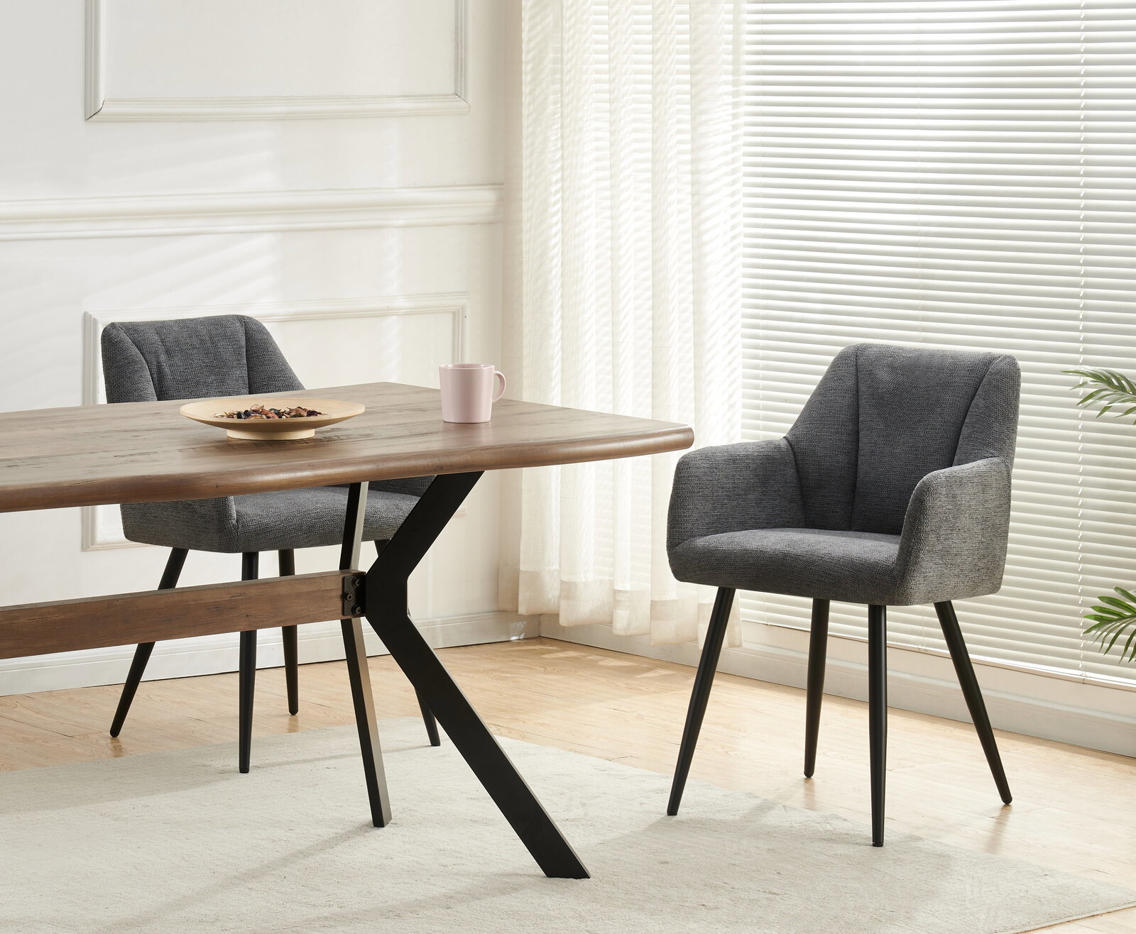 Schwarzer Esstisch mit Holzplatte und X-förmigem Metallgestell, daneben zwei graue Polsterstühle im hellen Esszimmer; Perspektive: seitliche 3/4-Ansicht auf Tisch und Stühle.