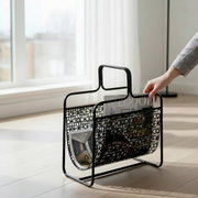 Schwarzer Metall-Zeitschriftenständer mit dekorativem Gittermuster und Tragegriff, gefüllt mit Magazinen, auf hellem Holzboden am Fenster; Perspektive: leicht seitliche Vorderansicht auf Bodenhöhe.