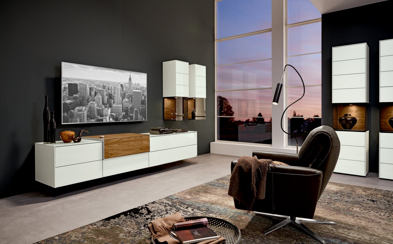 Modernes Wohnzimmer mit TV-Medienelement in Weiß und Holz, Wandregalen und einem großen Fenster mit Abendhimmel, seitliche Perspektive.