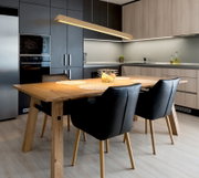 Moderner Essbereich in der Küche mit massivem Holztisch, vier schwarzen Polsterstühlen und länglicher Holz-Pendelleuchte, aufgenommen aus leicht schräger Frontperspektive auf Tisch und Stühle