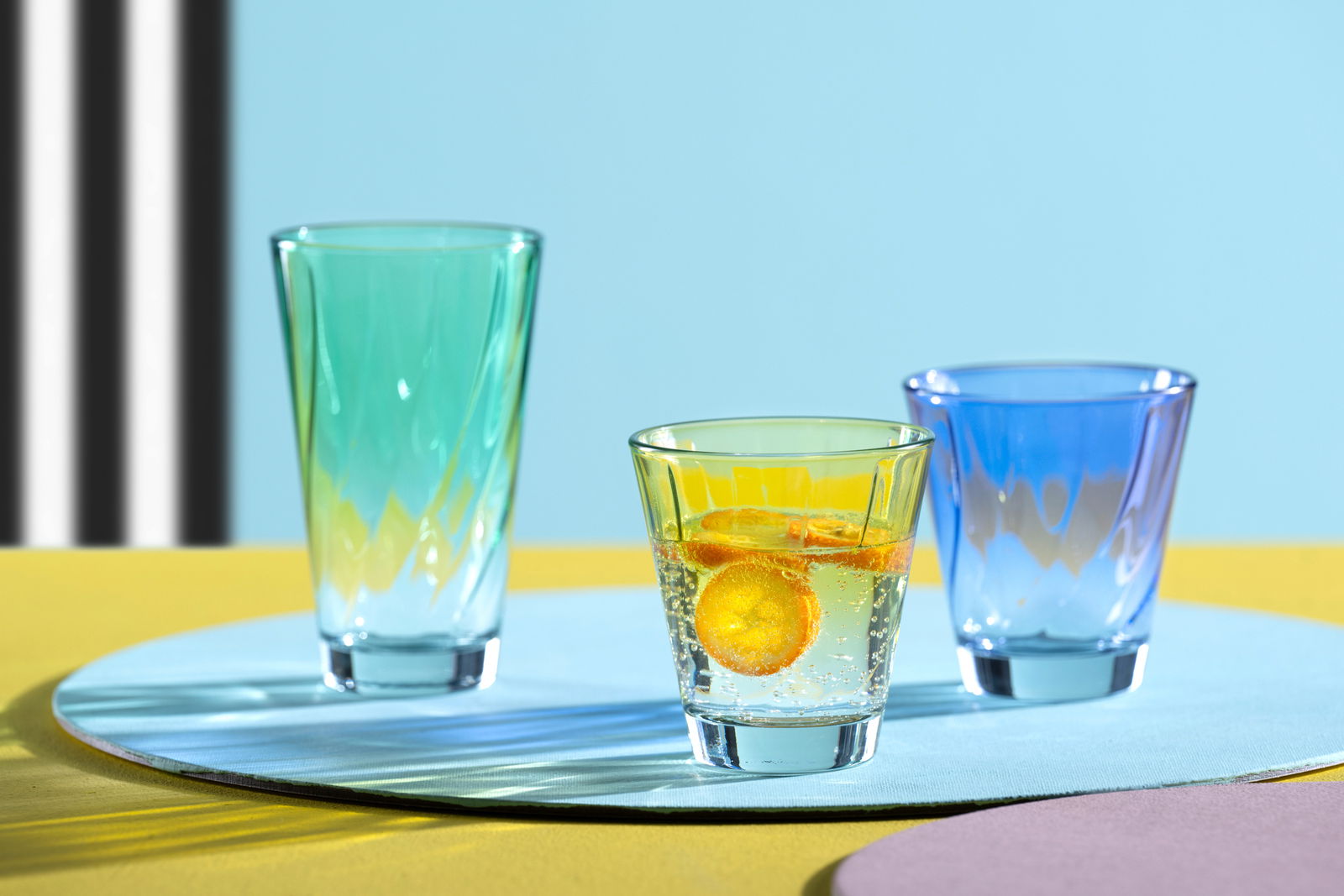 Leonardo Trinkglas TWIST Drei bunte Trinkgläser auf einem Tisch, mit einem blauen Glas im Vordergrund. Perspektive von vorne.