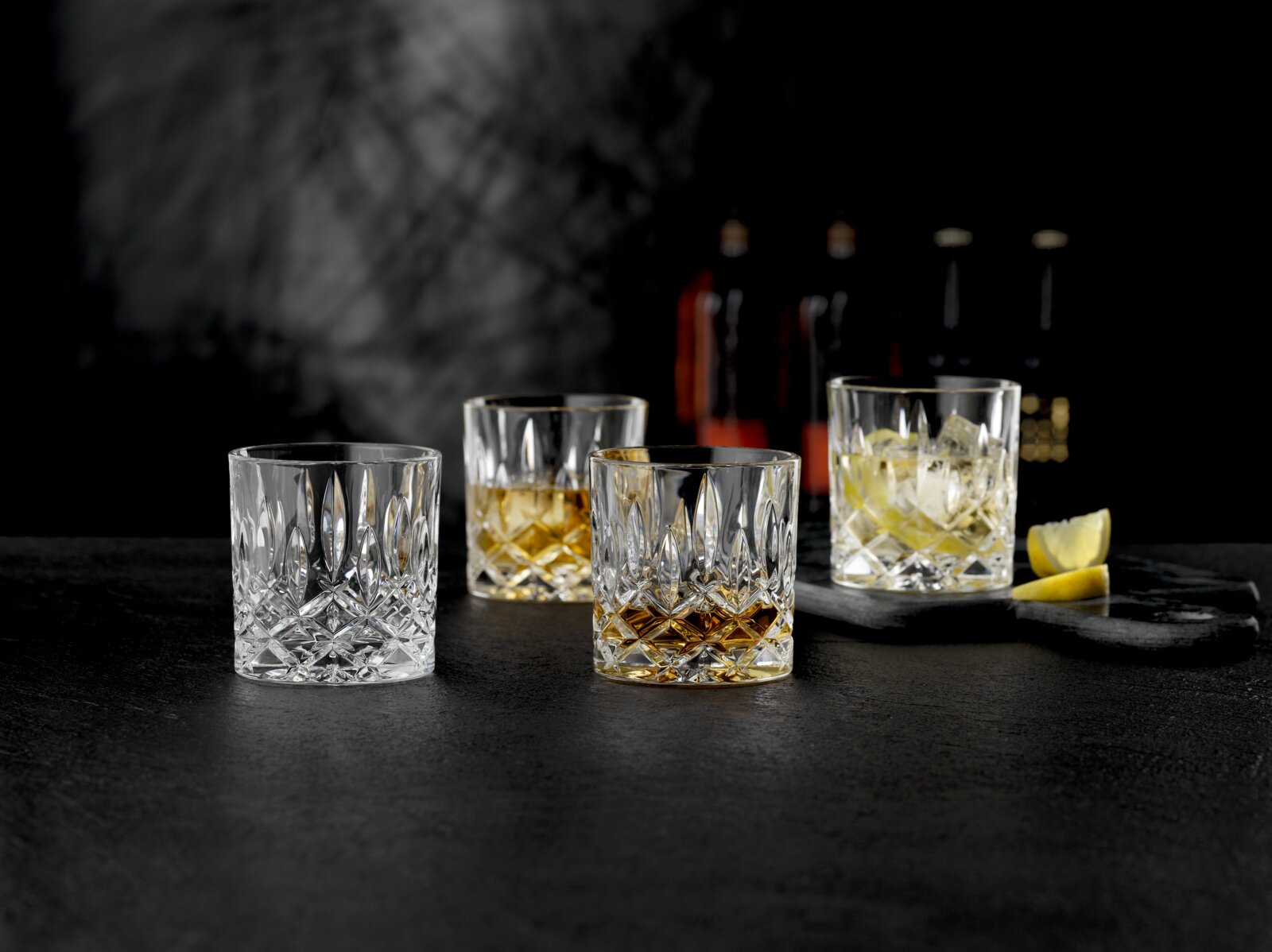 Nachtmann Single Old Fashioned Becher NOBLESSE Vier facettierte Kristall-Whiskygläser (Tumbler) auf dunkler Oberfläche, teils mit Whisky und Eis, im Vordergrund in leichter Seitenansicht aufgenommen; im Hintergrund unscharfe Flaschen und Zitronenspalten.