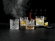 Vier facettierte Kristall-Whiskygläser (Tumbler) auf dunkler Oberfläche, teils mit Whisky und Eis, im Vordergrund in leichter Seitenansicht aufgenommen; im Hintergrund unscharfe Flaschen und Zitronenspalten.