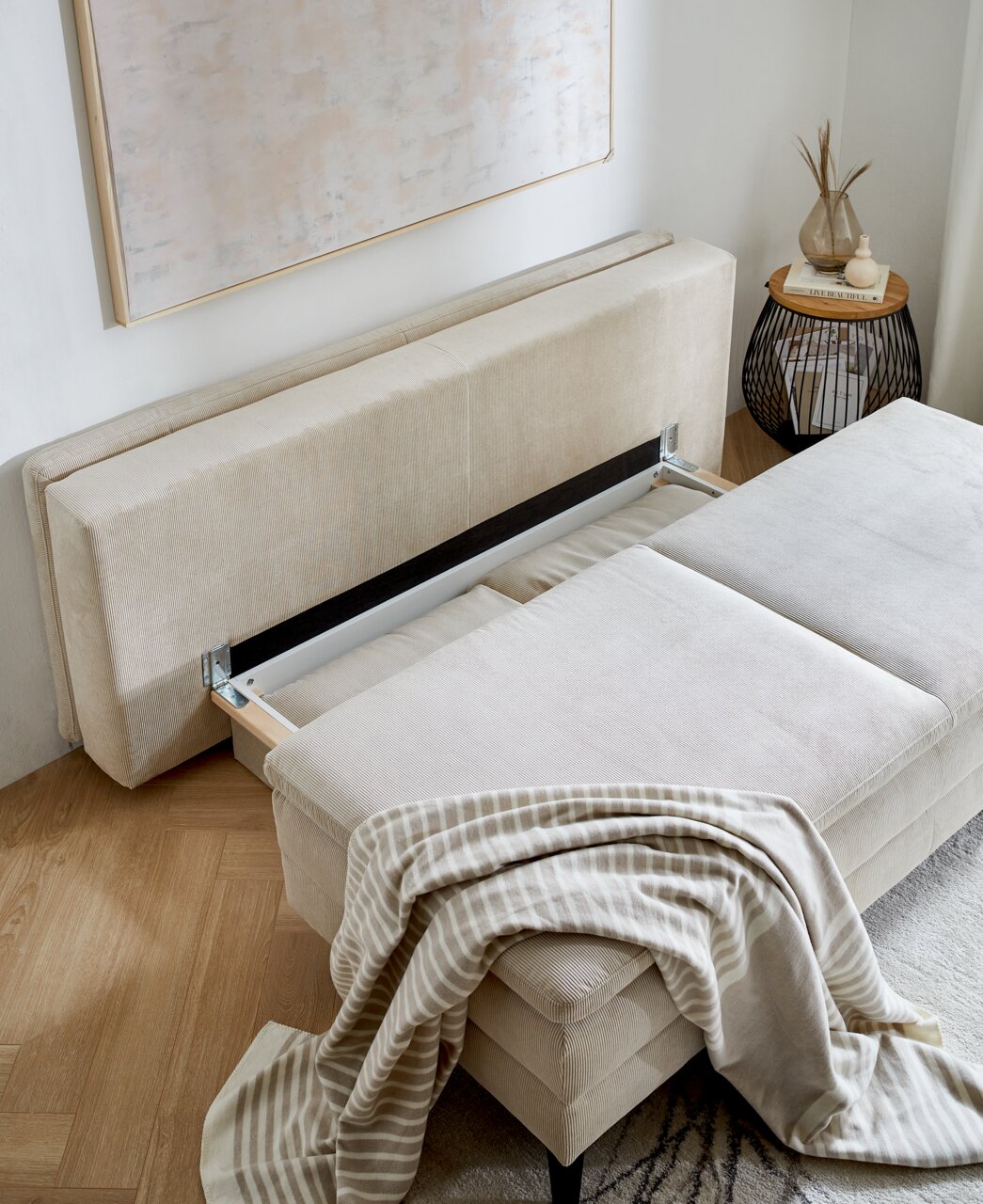 Querschläfer-Sofa mit Hubbeschlag in beiger Farbe, von oben fotografiert, mit einer gestreiften Decke darauf.