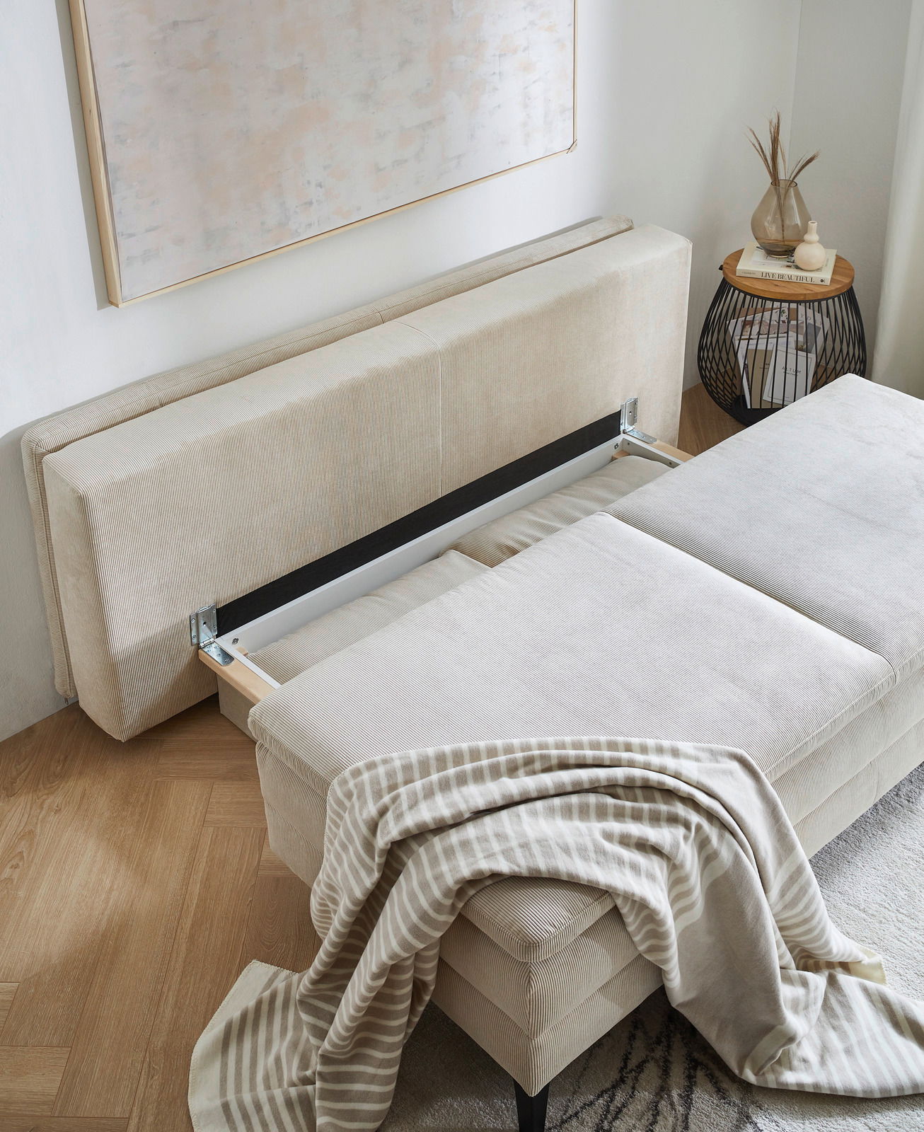 Querschläfer-Sofa mit Hubbeschlag in beiger Farbe, von oben fotografiert, mit einer gestreiften Decke darauf.