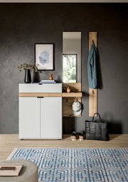 Modernes Flur-Set mit weißer Schuhkommode und Holzablage, Wandspiegel, Garderobenpaneel mit Jacke sowie Tasche und Schuhen auf dem Boden, frontal aus Augenhöhe fotografiert.