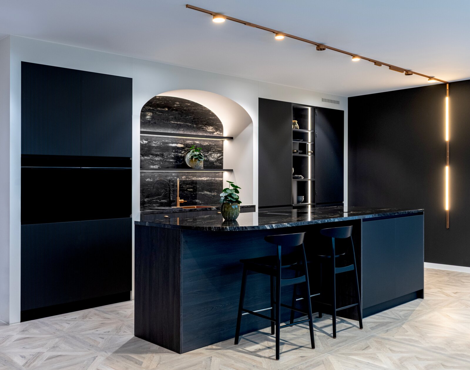 Moderne Designküche in Dunkelblau/Schwarz mit Kücheninsel und dunkler Stein-Arbeitsplatte, zwei Barhockern, Nischenrückwand in Steinoptik und Decken-Schienenbeleuchtung, aufgenommen aus schräger Frontansicht