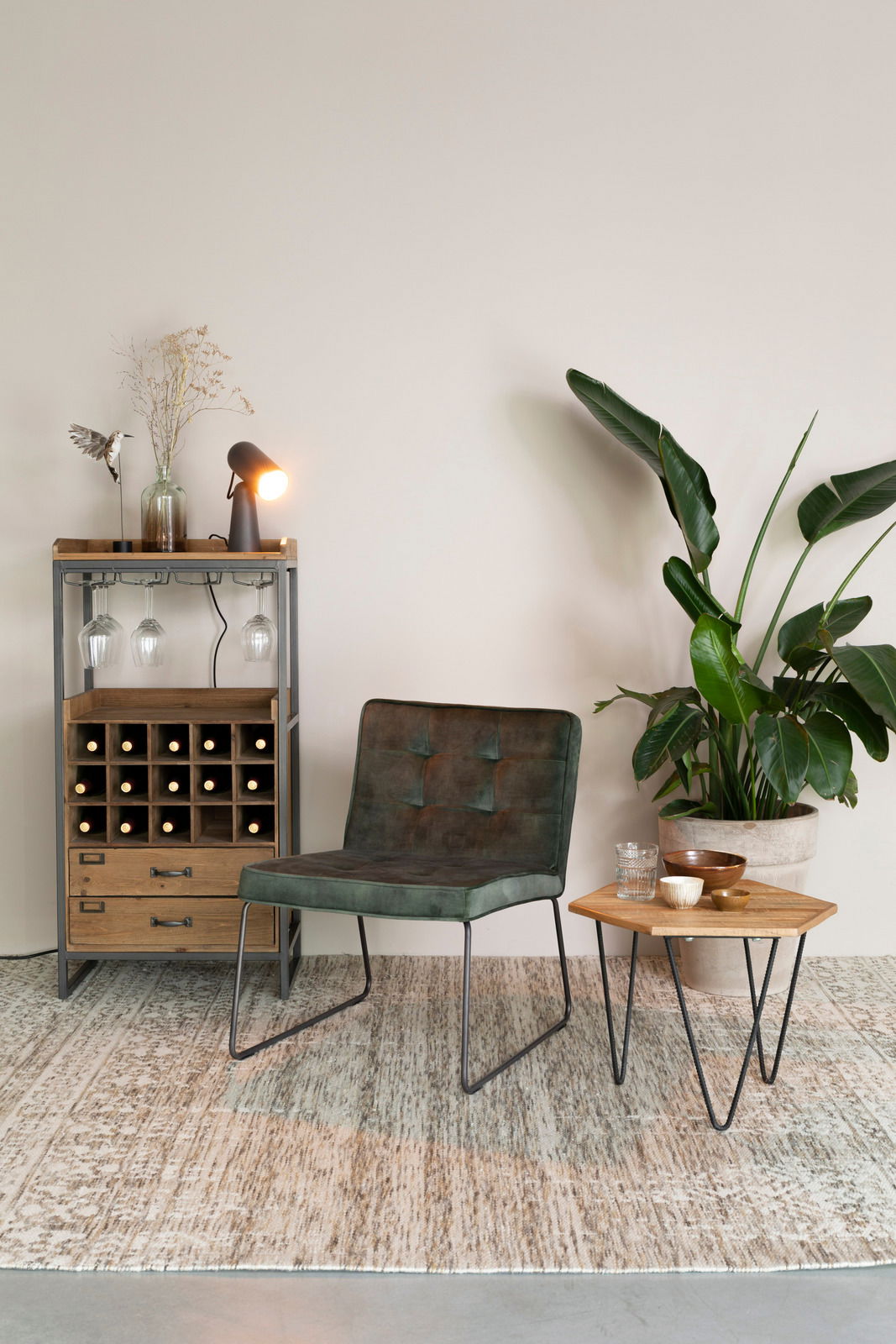 Weinregal aus Holz und Metall mit integrierten Glashaltern, dekoriert mit einer Lampe und einer Vase, im Vordergrund ein grüner Sessel und ein kleiner Tisch mit Pflanzen im Hintergrund, seitliche Perspektive.