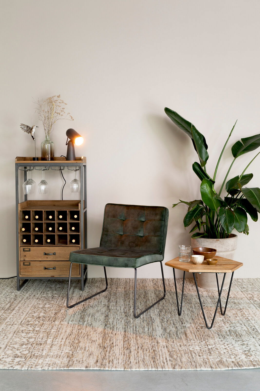 Weinregal aus Holz und Metall mit integrierten Glashaltern, dekoriert mit einer Lampe und einer Vase, im Vordergrund ein grüner Sessel und ein kleiner Tisch mit Pflanzen im Hintergrund, seitliche Perspektive.