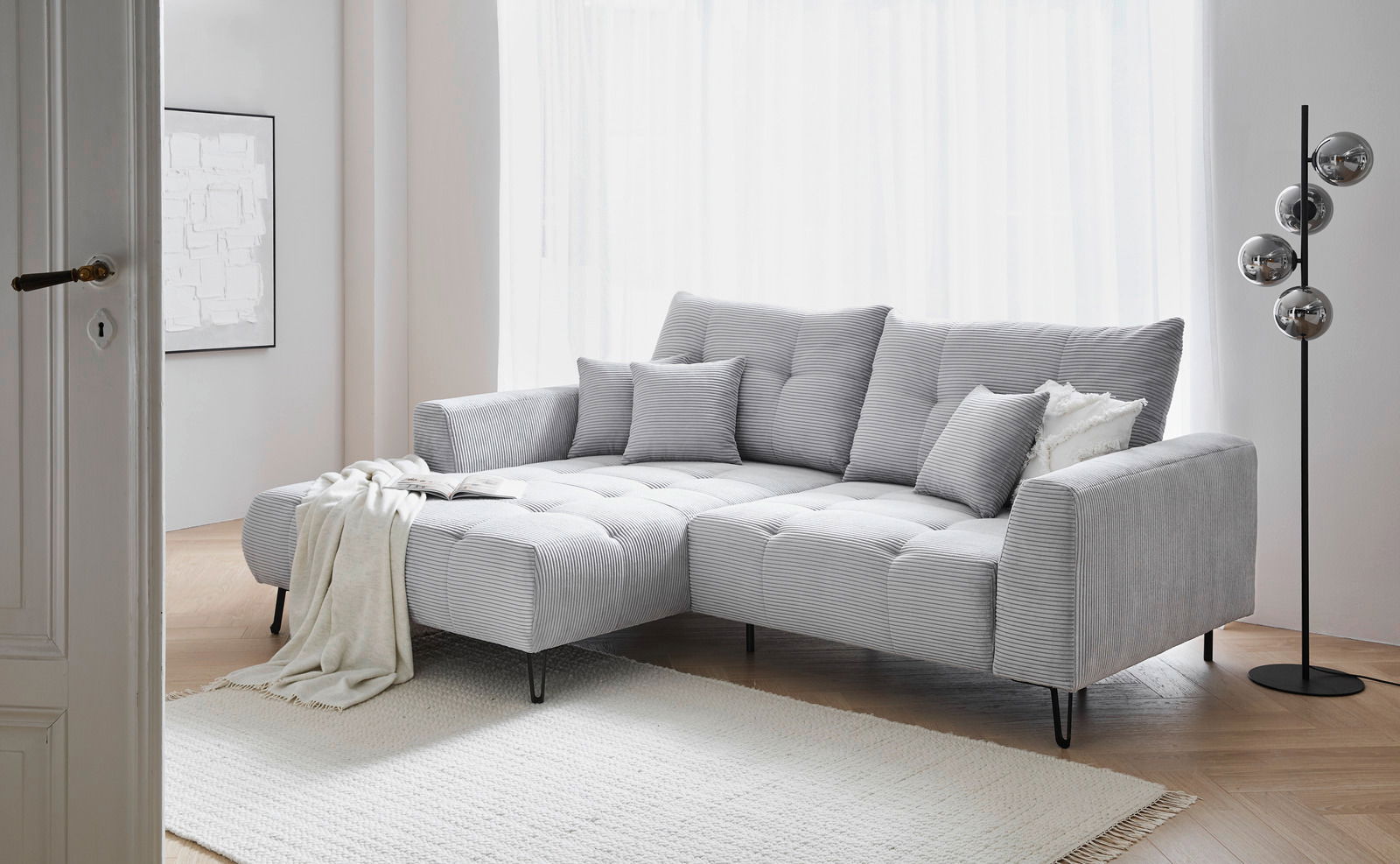 Graues Ecksofa mit Ottomane links, dekoriert mit Kissen und Decke, in einem hellen Wohnzimmer mit Holzboden und Stehlampe, seitliche Perspektive.