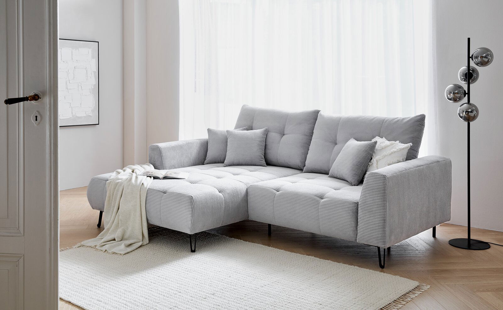 Graues Ecksofa mit Ottomane links, dekoriert mit Kissen und Decke, in einem hellen Wohnzimmer mit Holzboden und Stehlampe, seitliche Perspektive.
