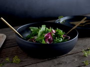 Schwarze, matte Salatschüssel mit frischem Blattsalat und goldfarbenem Besteck auf rustikalem Holztisch, aufgenommen in schräger Seitenansicht von vorne