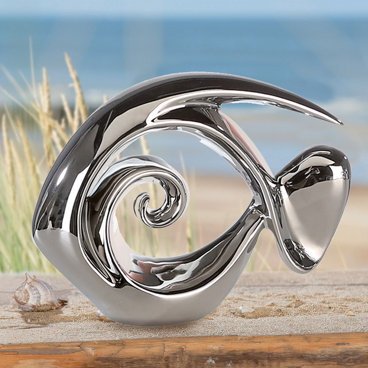 Silberne Fischskulptur in moderner Form, seitliche Perspektive, auf einem Tisch vor einem Strandhintergrund.