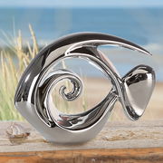 Silberne Fischskulptur in moderner Form, seitliche Perspektive, auf einem Tisch vor einem Strandhintergrund.